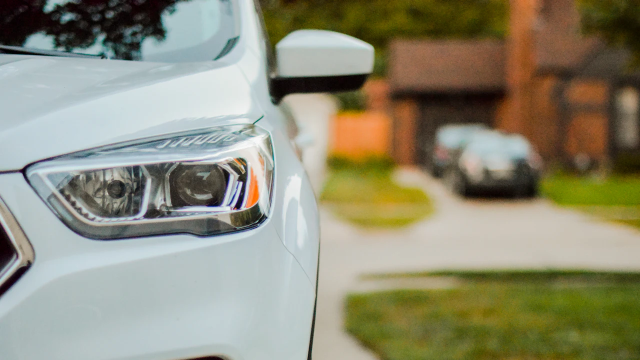 The front corner and headlight of a white SUV parked on a driveway, with a house and another parked car out of focus behind