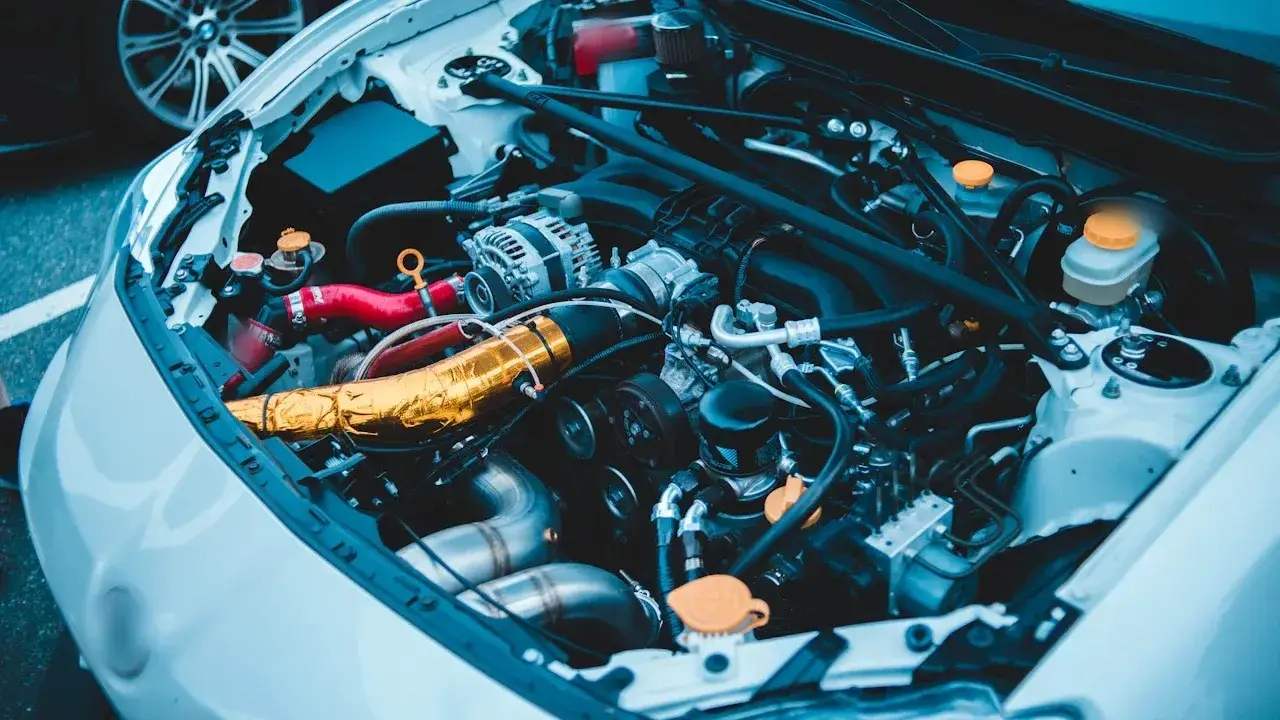 An open engine bay on a white car: the alternator, red silicone hoses, a foil-wrapped intake pipe, the oil filter and fluid reservoirs with orange caps.
