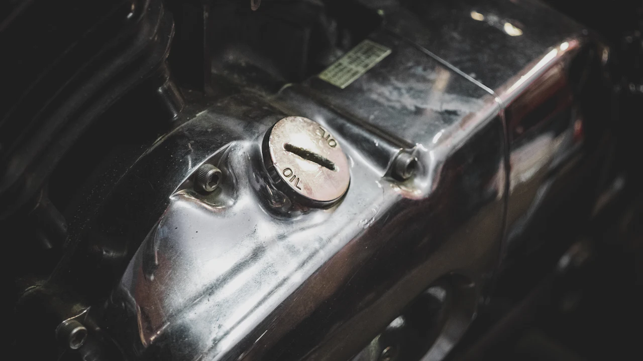 A close-up of an engine's metal rocker cover in low light, with a round oil filler cap marked OIL, a small printed label and bolts along the cover's edge.