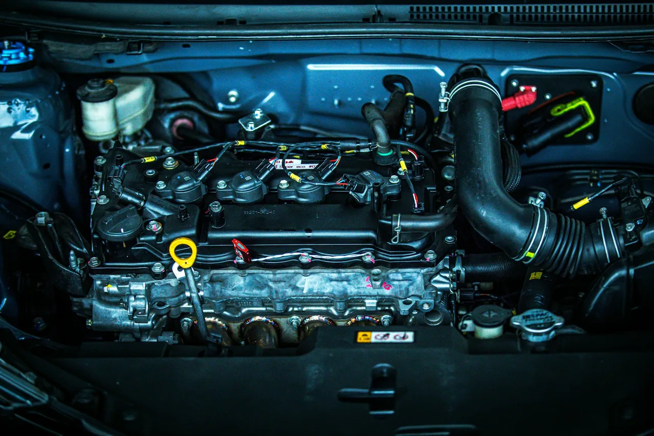 An open engine bay under blue-toned light: a four-cylinder engine with its ignition coils in a row on top, a large black intake hose on the right, a coolant reservoir at the top left and a yellow oil dipstick handle.