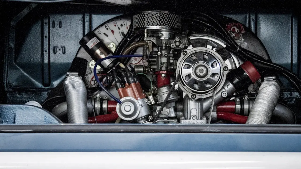 The engine bay of an older air-cooled car, with carburettor, cooling fan housing and red hoses, decals blurred.