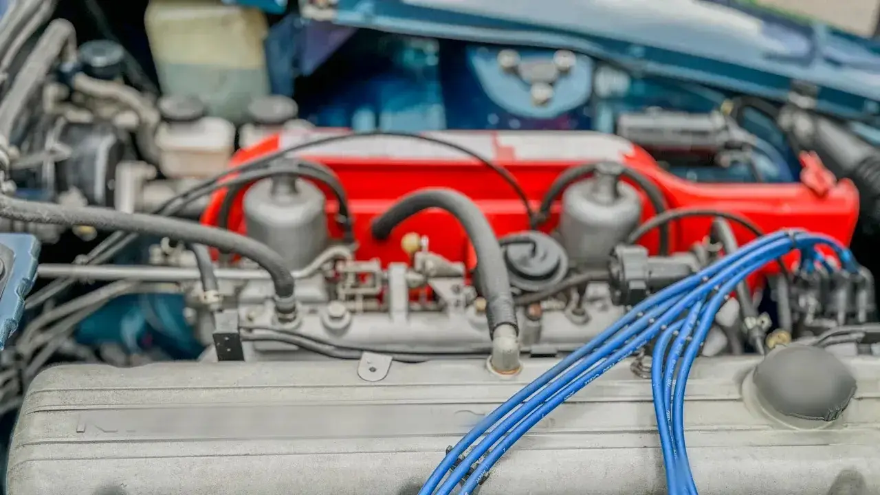 An older four-cylinder engine under the hood: a finned valve cover, blue spark plug leads, a red intake with twin carburettors, hoses and the oil filler cap.
