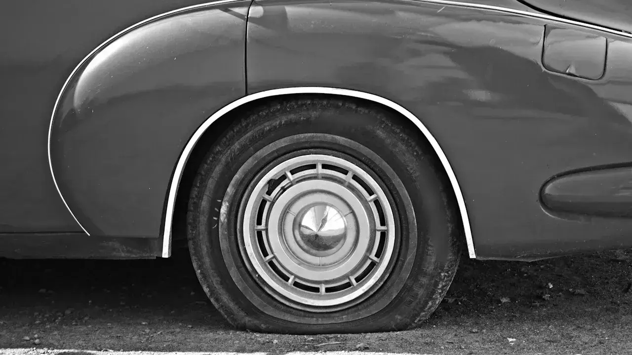 Black-and-white close-up of an older car's rear wheel arch, with a flat tyre, a chrome hubcap and the curved bodywork above.