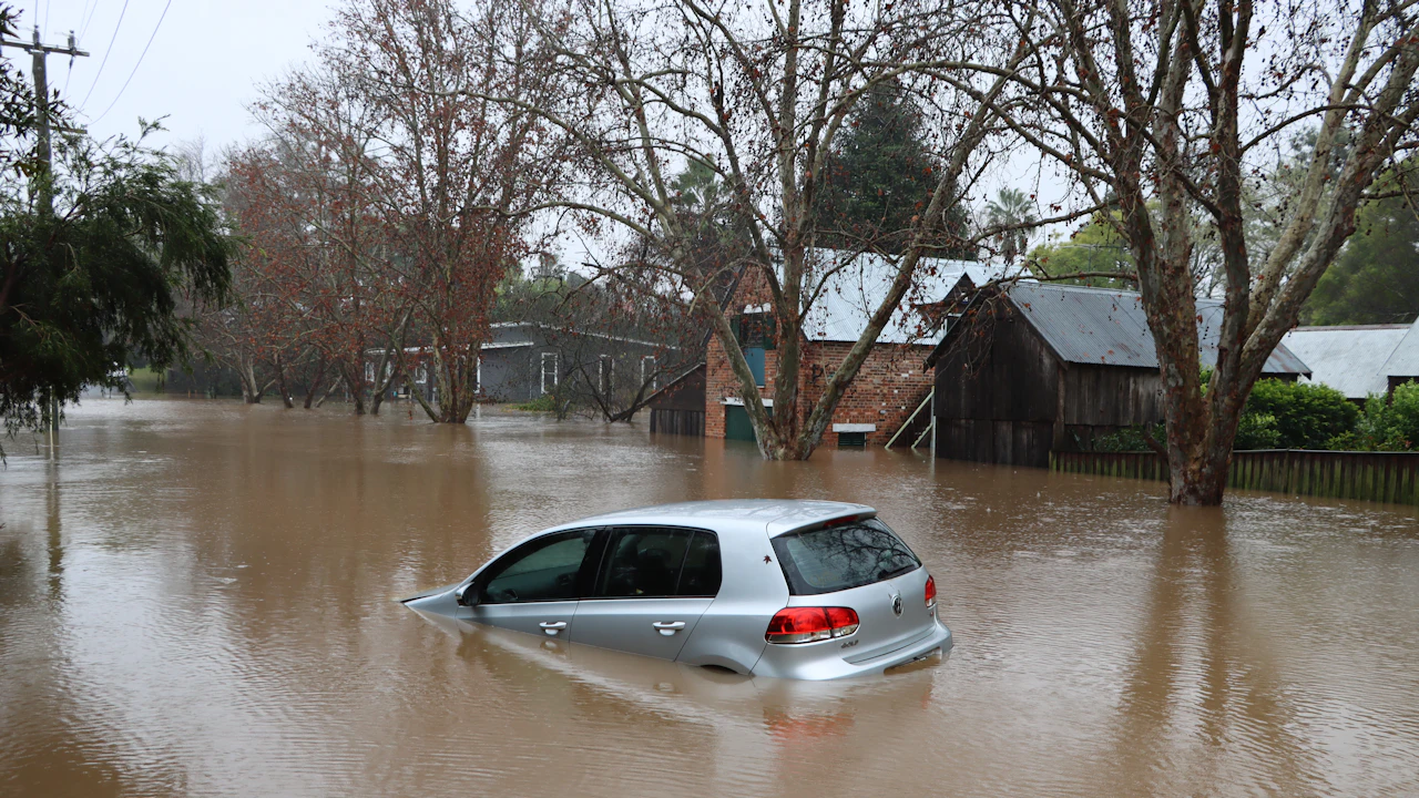 A silver hatchback submerged to the window line in brown floodwater on a residential street