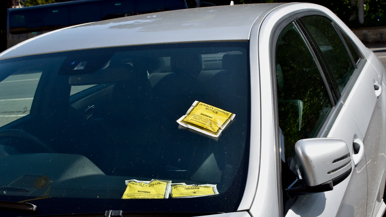 A dusty silver saloon parked at a kerb with three yellow penalty charge notices stuck to the outside of its windscreen, one high on the glass and two lower down, the wing mirror and door glass dark beside them