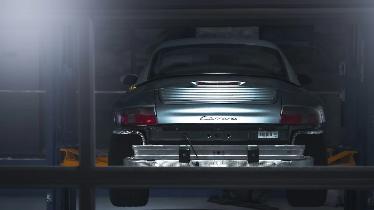 A silver sports car raised on a two-post lift in a dim workshop, seen from behind with the rear structure exposed