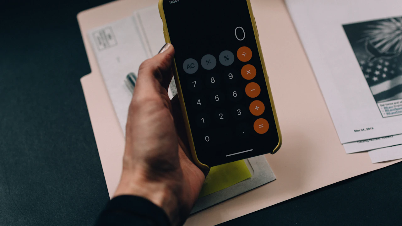 A hand holding a phone with the calculator app open, resting over a folder of printed documents on a dark desk
