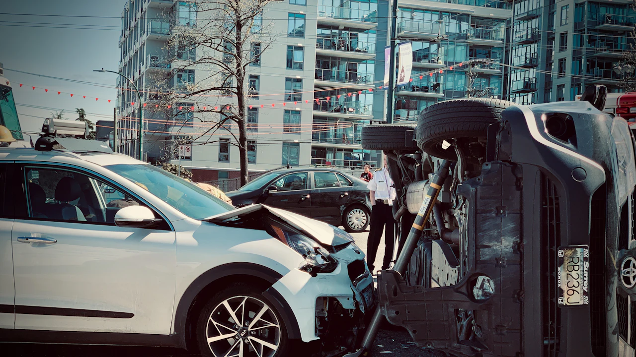 A multi-car collision on an urban street: a white SUV with a crushed front end, a second damaged car, and an SUV rolled onto its side, a person standing among the wreckage with apartment buildings behind.
