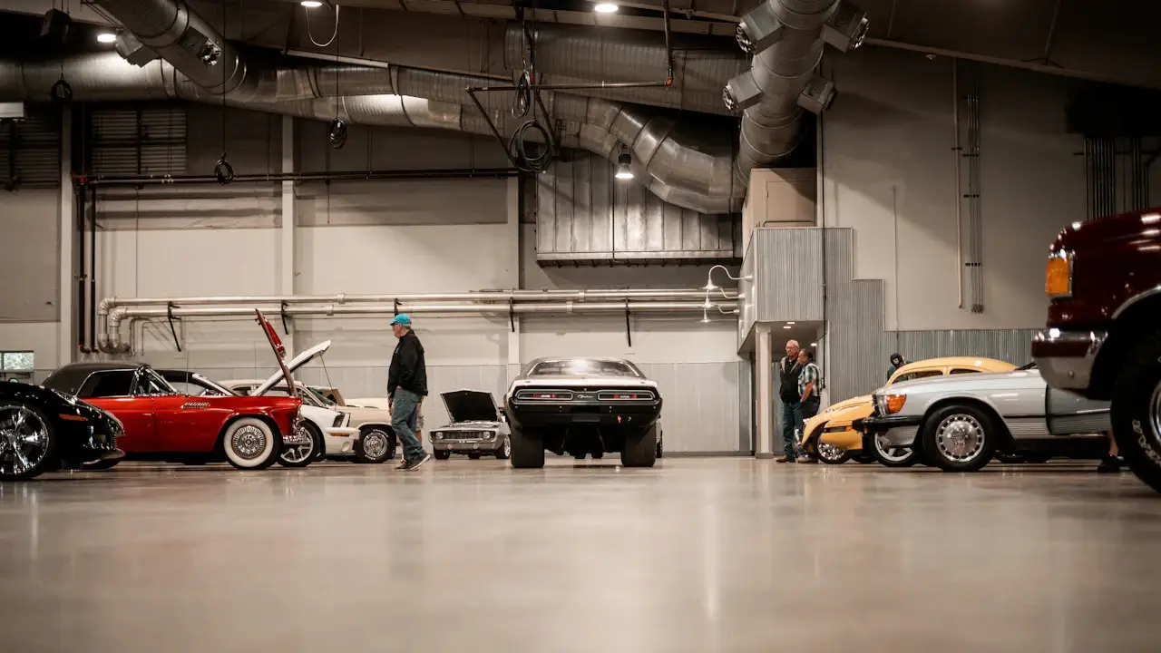 Used and classic cars parked in a large indoor hall, some with hoods raised, as a man walks between them.