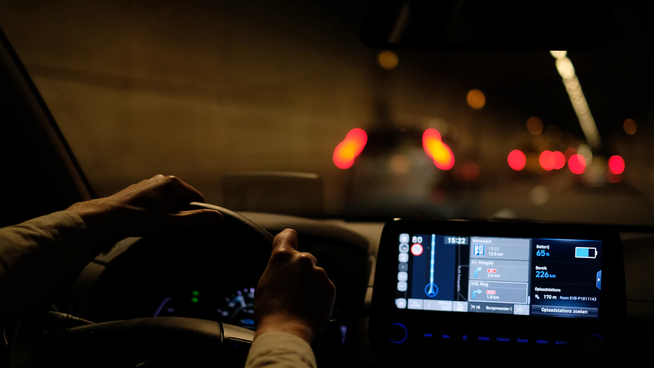 Hands on a steering wheel at night, dashboard glowing, blurred tail-lights through the windscreen ahead