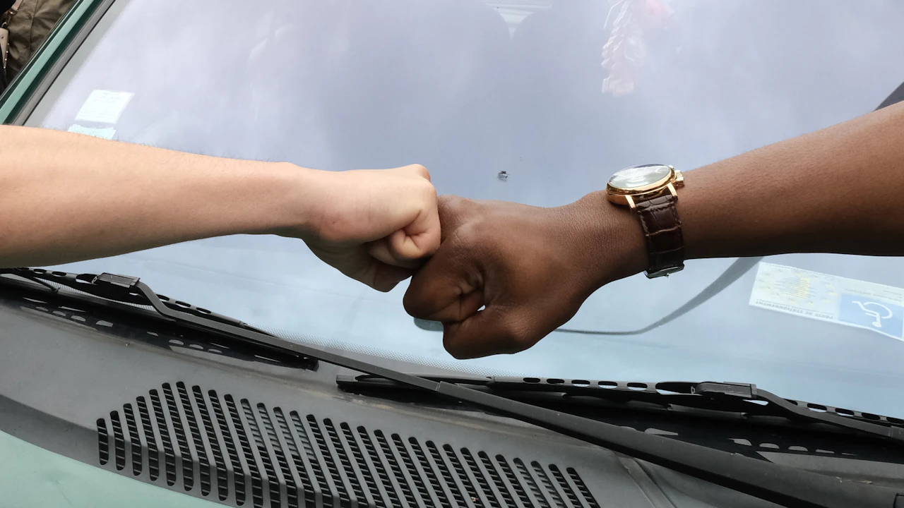 Two people bumping fists across the bonnet of a car, with the windscreen and wiper blades filling the lower half of the frame