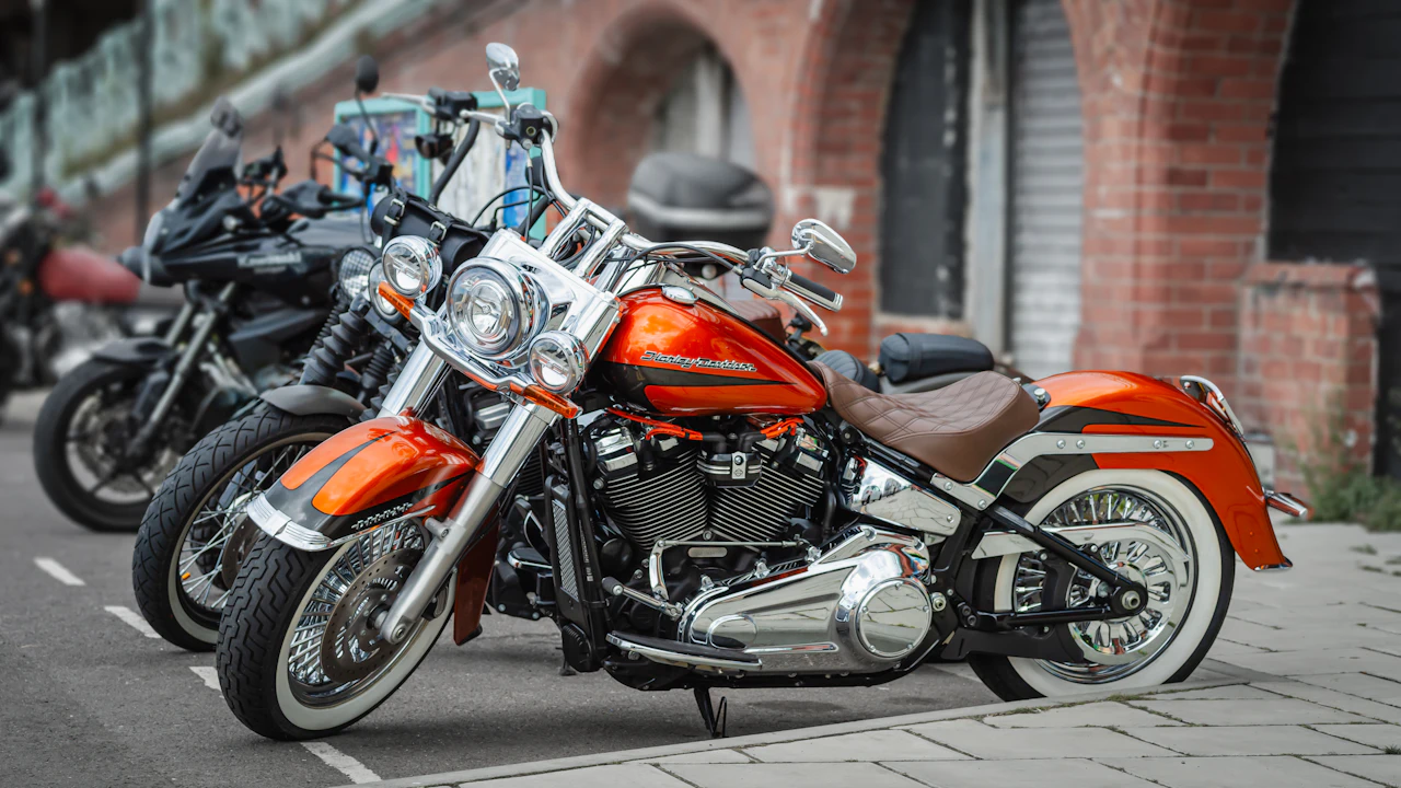 An orange Harley-Davidson cruiser with whitewall tyres parked in a row of motorcycles beside a brick arcade