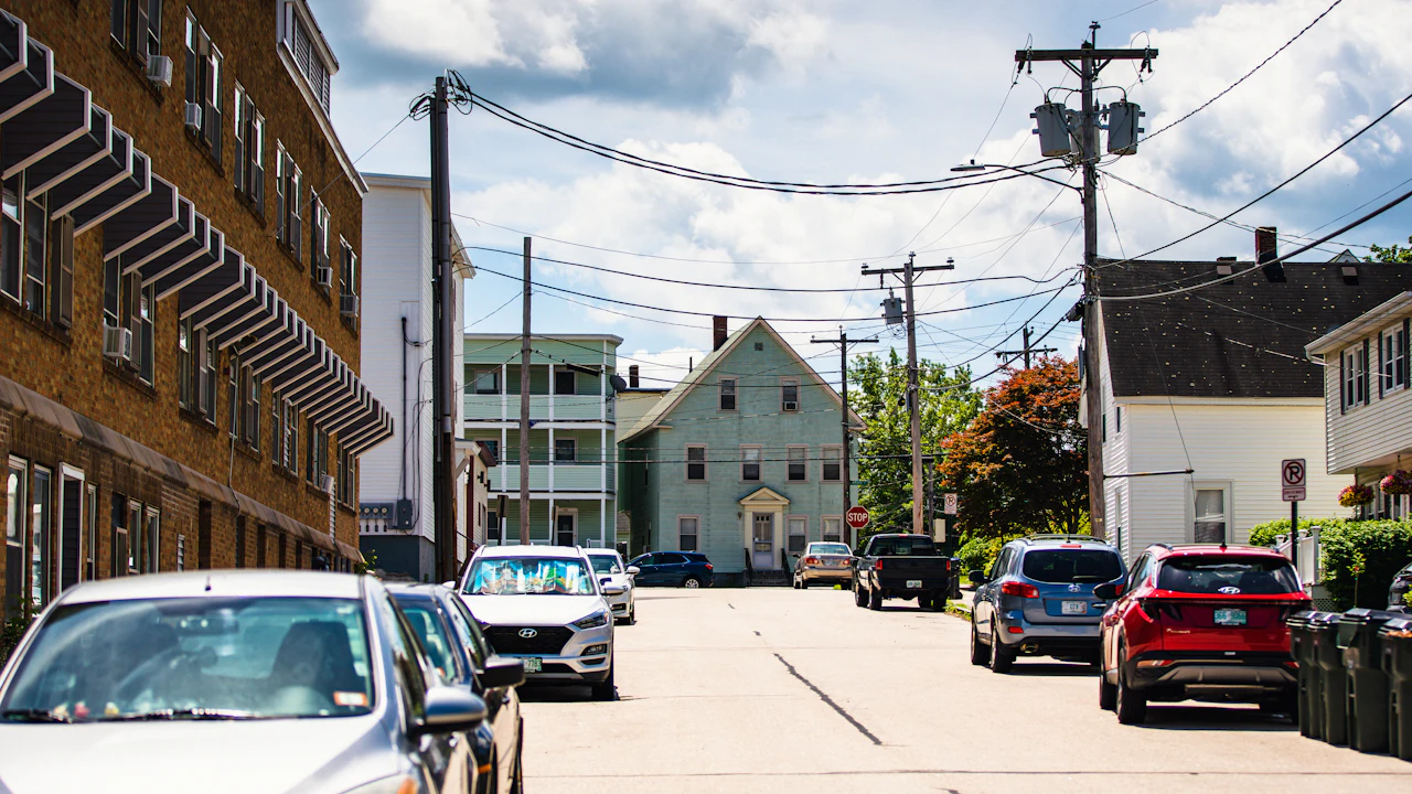 A New England side street lined with parked cars and clapboard triple-deckers, power lines overhead