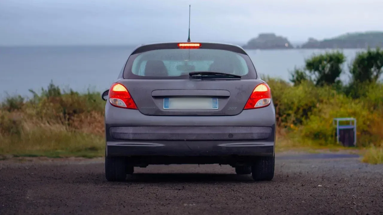 The rear of a grey hatchback parked on gravel above the sea, brake lights on, with its rear window, rear wiper and painted tailgate in view.