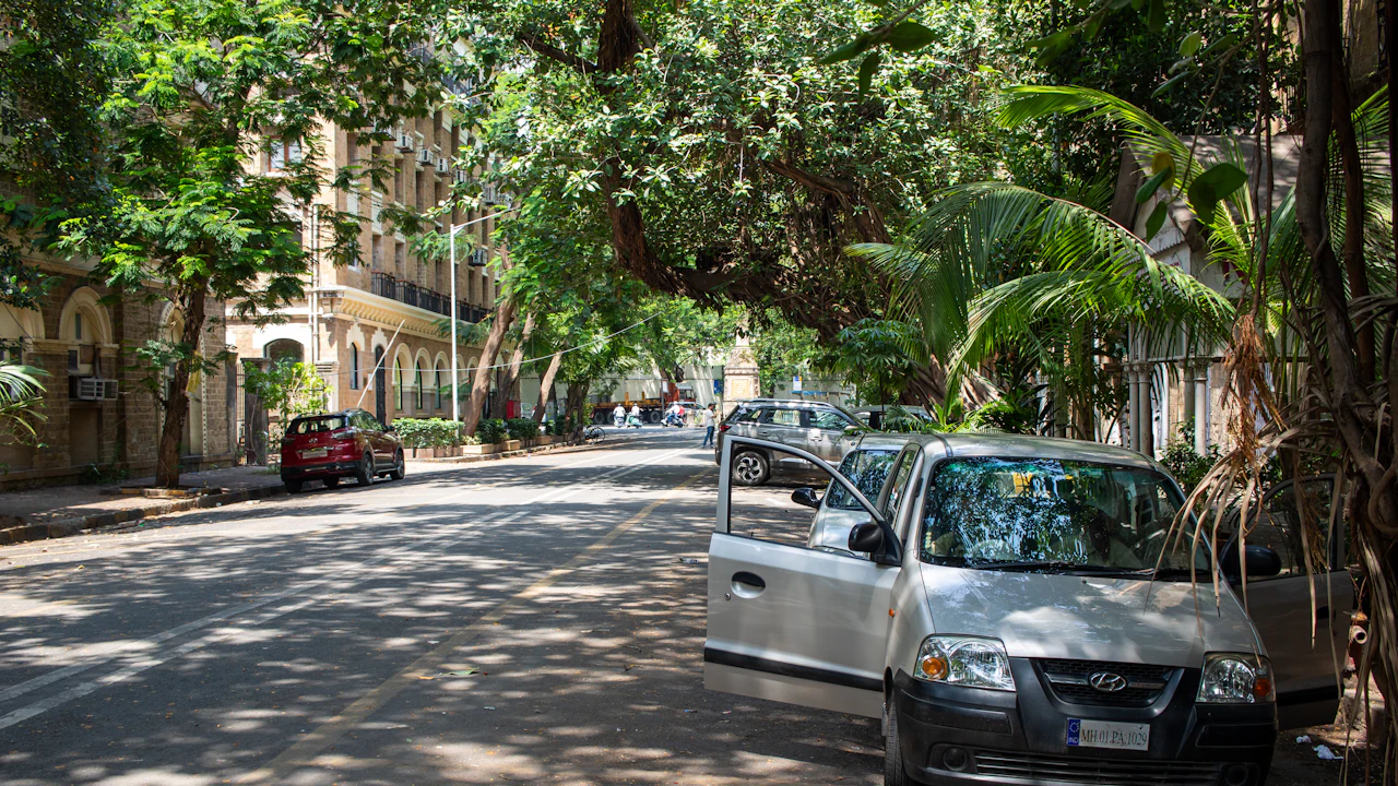A silver hatchback parked at the kerb of a quiet tree-lined street, one door standing open, tall palms and older apartment blocks behind it