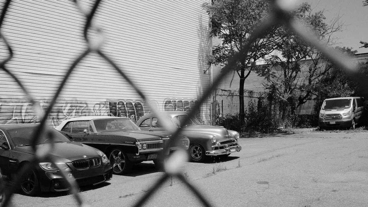 A black-and-white view through a chain-link fence of several cars parked in a lot beside a graffitied wall, including an older sedan and a van