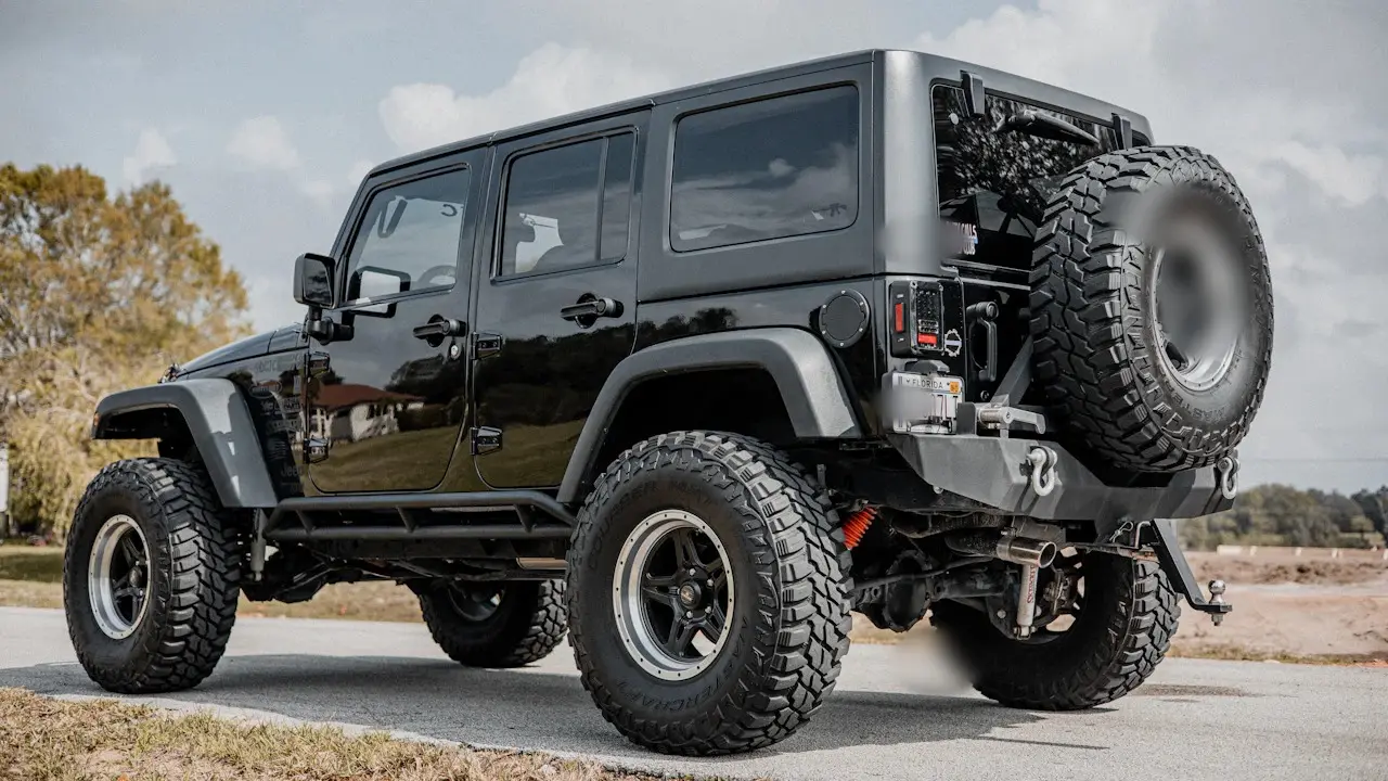 A lifted black Jeep seen from the rear quarter, with large tires, a rear bumper and a spare tire carrier.
