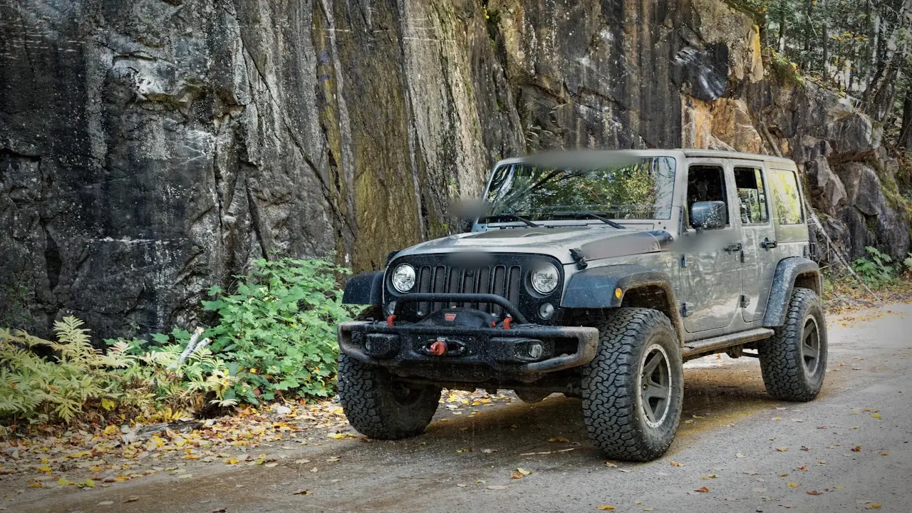 A grey Jeep with a front winch bumper parked on a dirt road below a rock face.