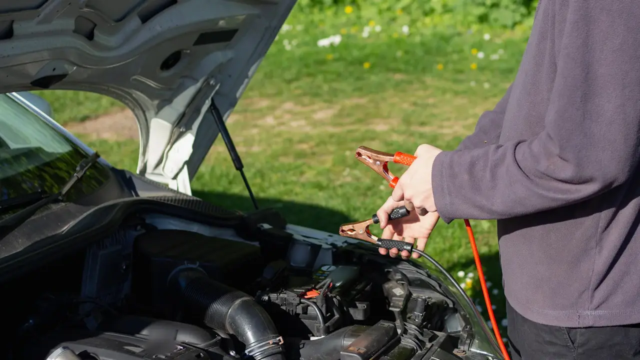 A person holding jumper-cable clamps beside a car with its hood open.