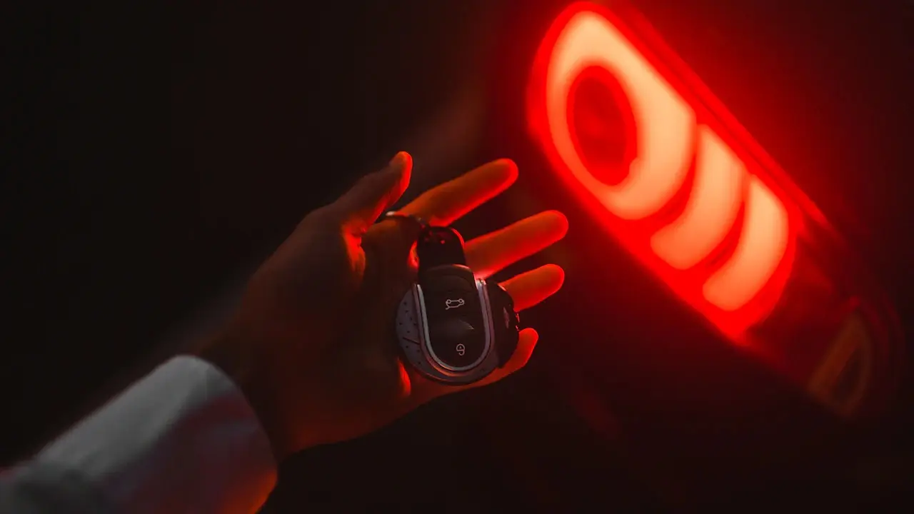 A hand holding a car key fob in front of a glowing red tail light.
