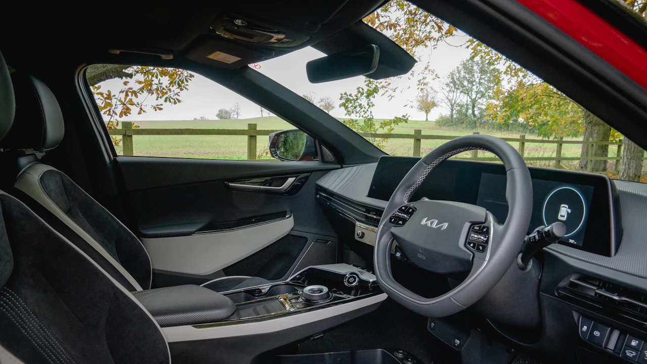 The interior of a modern Kia crossover photographed from the passenger side, twin screens across the dash and a fenced green field beyond the windows
