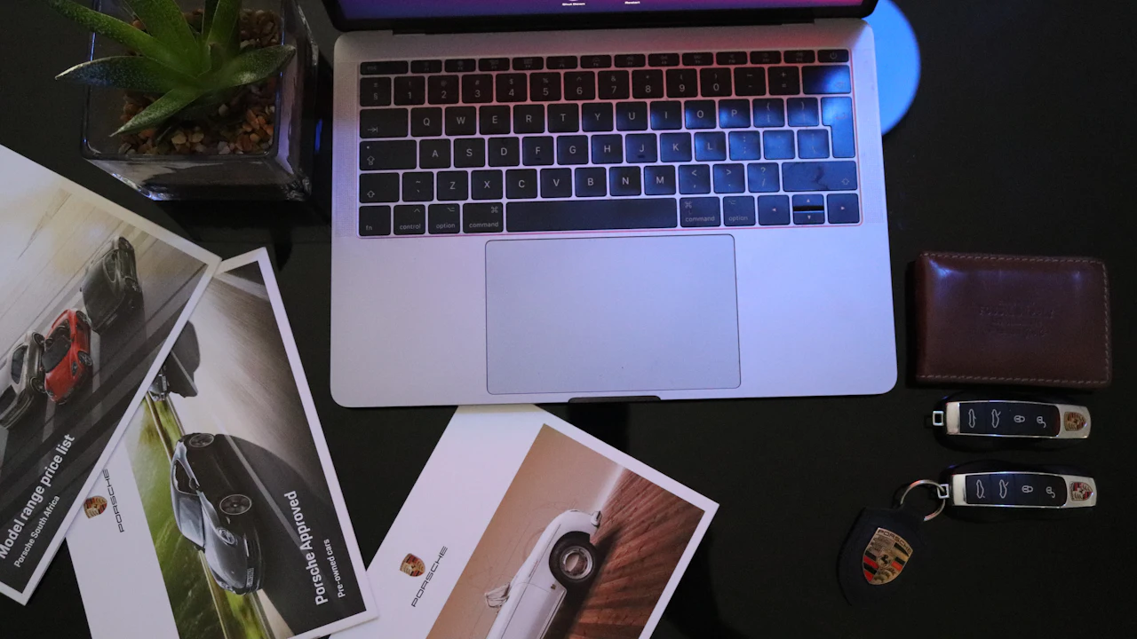 An overhead view of a laptop on a dark desk surrounded by manufacturer brochures, a wallet and two car key fobs
