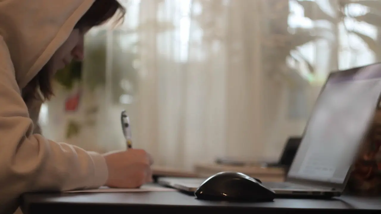 A person writing on paper beside an open laptop at a desk.