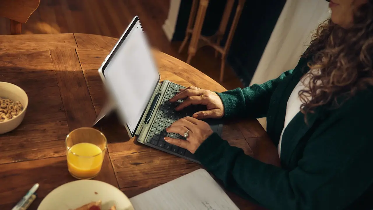 A person typing on a tablet keyboard at a wooden kitchen table, a glass of juice beside it; the screen's text blurred.