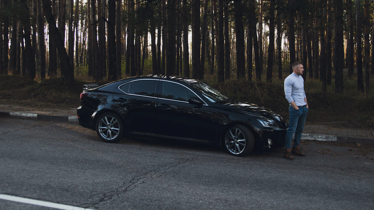 A black Lexus sedan parked on a forest road at dusk, a man standing beside the passenger door