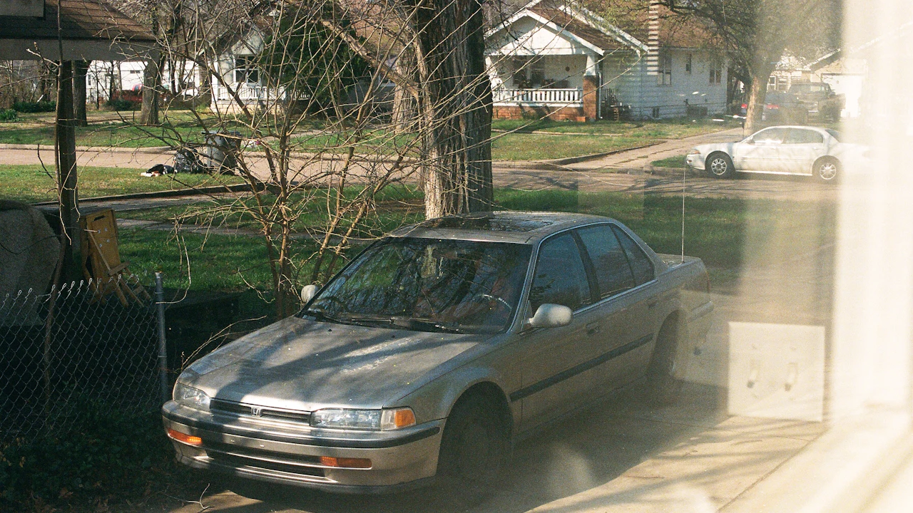 A silver early-1990s sedan sitting in a suburban driveway, photographed through a window, with houses along the street beyond