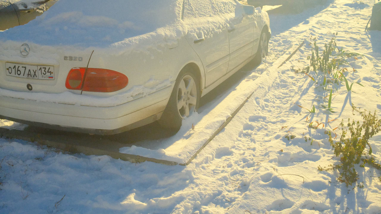 A silver Mercedes saloon parked outdoors under a thick covering of fresh snow, low winter sun catching the roofline and rear wing