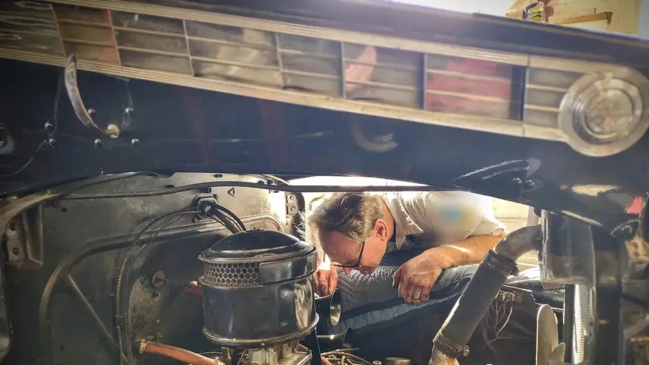 A mechanic leaning in under the raised hood of an older car, beside the round air cleaner, hoses and wiring of the engine bay.