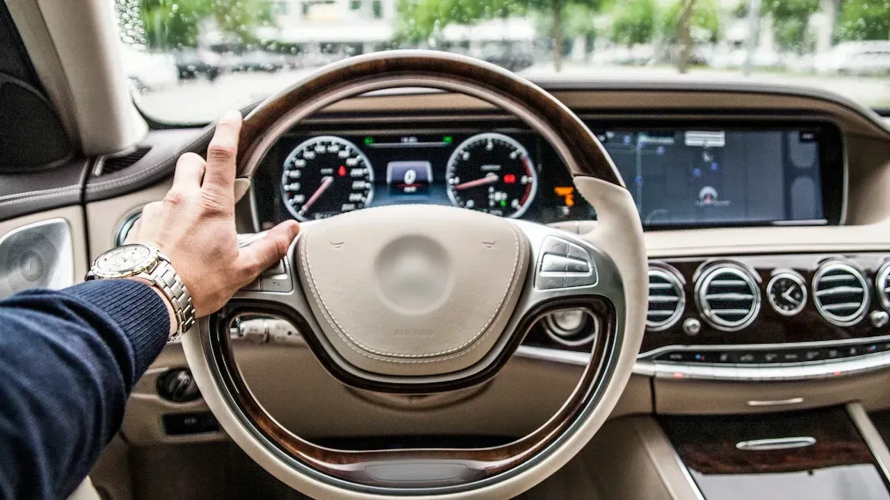 A driver's hand on the wood-rimmed steering wheel of a luxury sedan with a cream leather dashboard, a wide instrument display with two round dials, a navigation screen to its right, a row of round chrome air vents with a small analogue clock between them, and a windscreen looking out on trees.