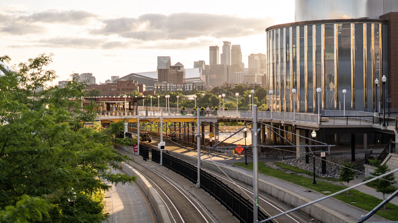 A Minneapolis cityscape at dusk, light-rail tracks curving below a glass building and the downtown skyline beyond