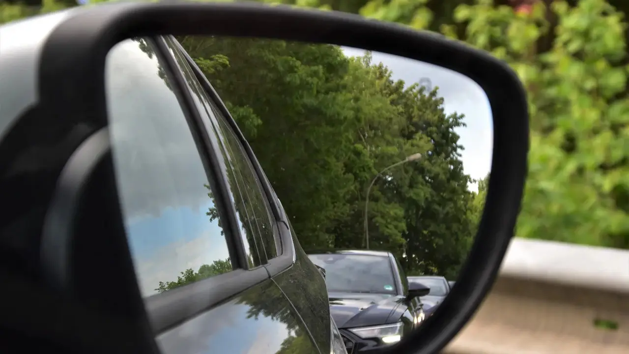 A car's side mirror reflecting trees and the cars behind.