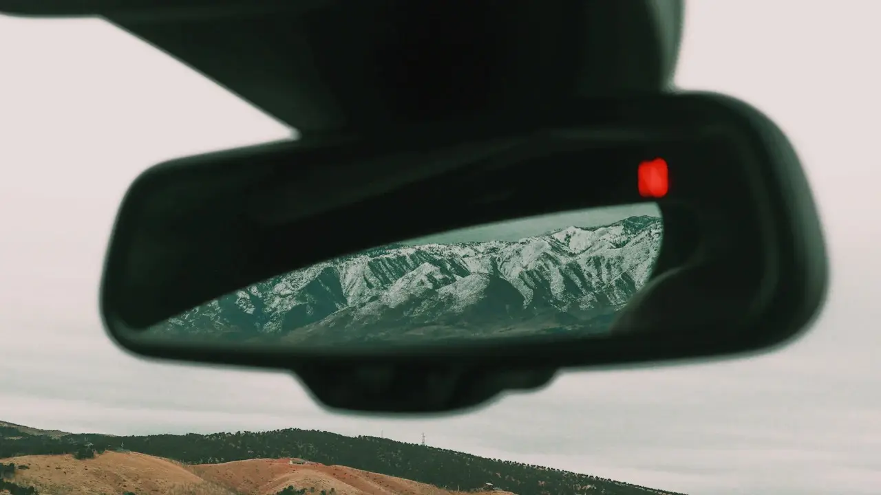 A car's rear-view mirror reflecting snow-capped mountains, a dry hillside below.