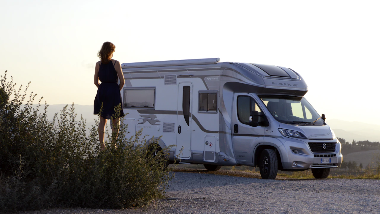 A silver coachbuilt motorhome parked on gravel at sunset, a woman standing beside it looking out across dry hills