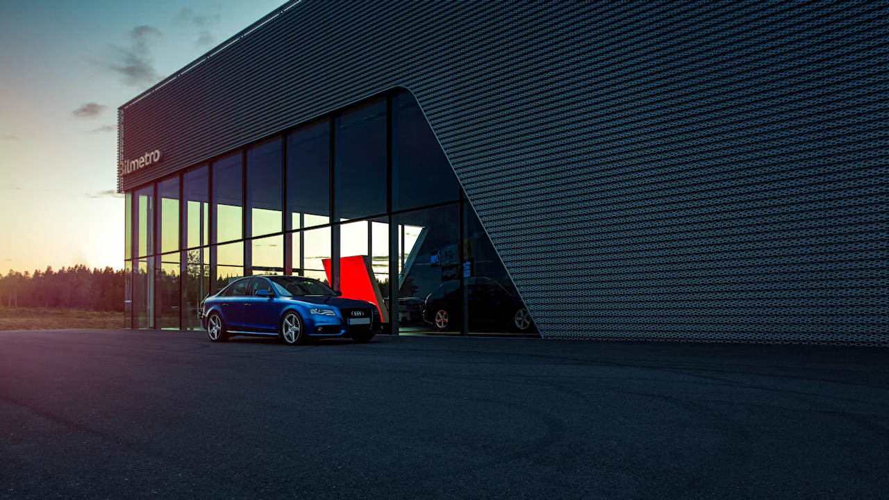 A blue saloon parked outside a modern glass-fronted dealership at dusk, more cars visible through the lit showroom windows