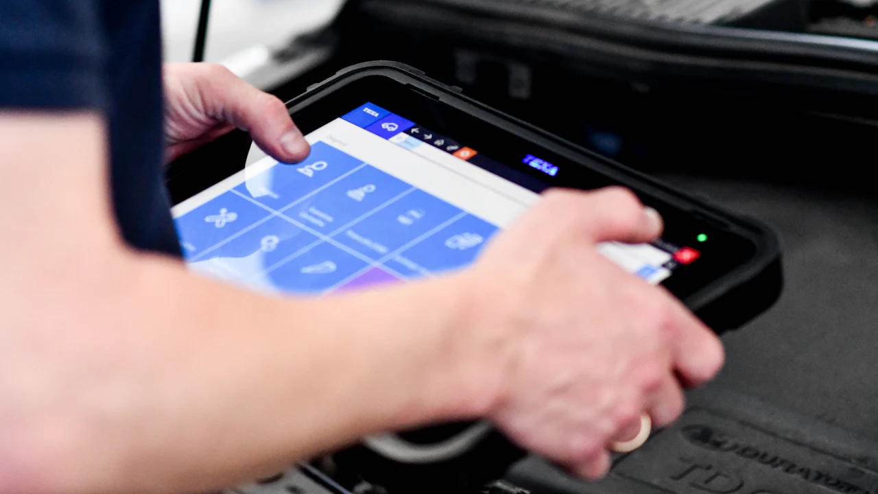 A technician holding a diagnostic scan tablet over the open engine bay of a car