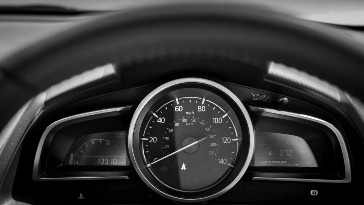 A black-and-white close-up of a car instrument cluster seen through the steering wheel: a large central speedometer marked in mph and km/h, a tachometer and odometer on the left reading 13910 miles, and a fuel and range display on the right.
