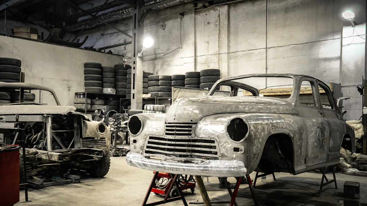 A stripped car body shell in bare metal standing on axle stands inside a workshop lined with stacked tyres