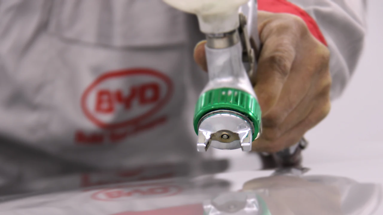 A spray gun held close over a car panel in a workshop, the paint laying down in a wet edge, the operator&rsquo;s sleeve blurred behind