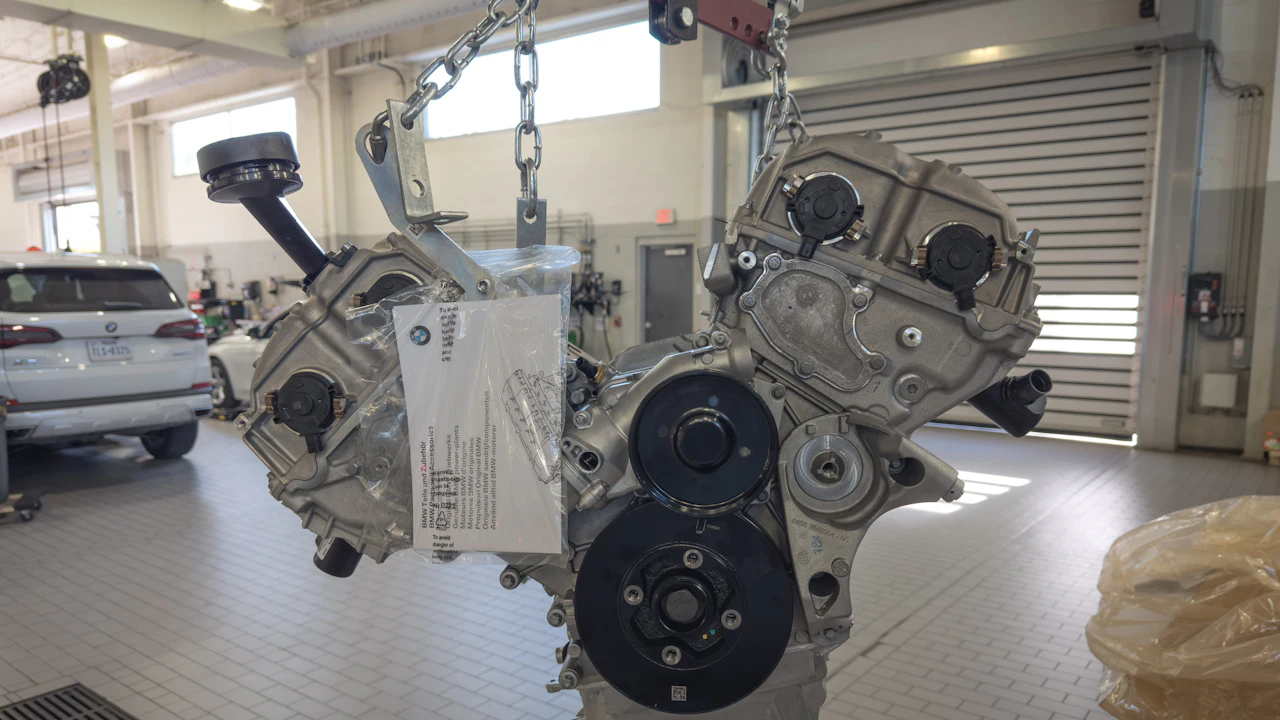 A bare engine hanging from a chain hoist in a dealership workshop, a parts tag taped to its side and a white SUV behind