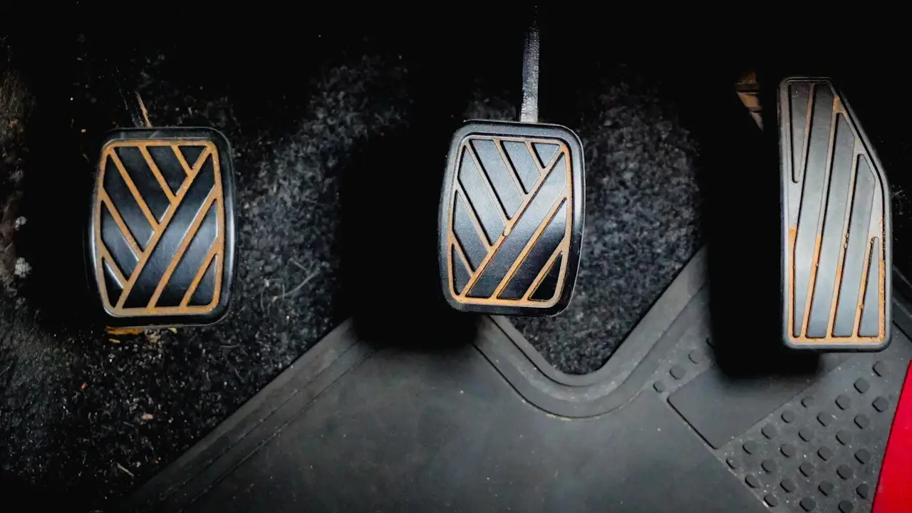 Three car pedals above a ridged rubber floor liner and carpet.