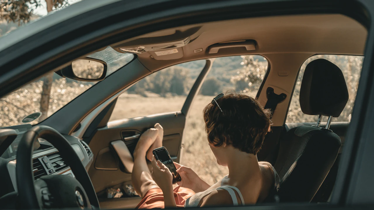 A person in the driver&rsquo;s seat of a parked car, seen through the open window, reading a phone