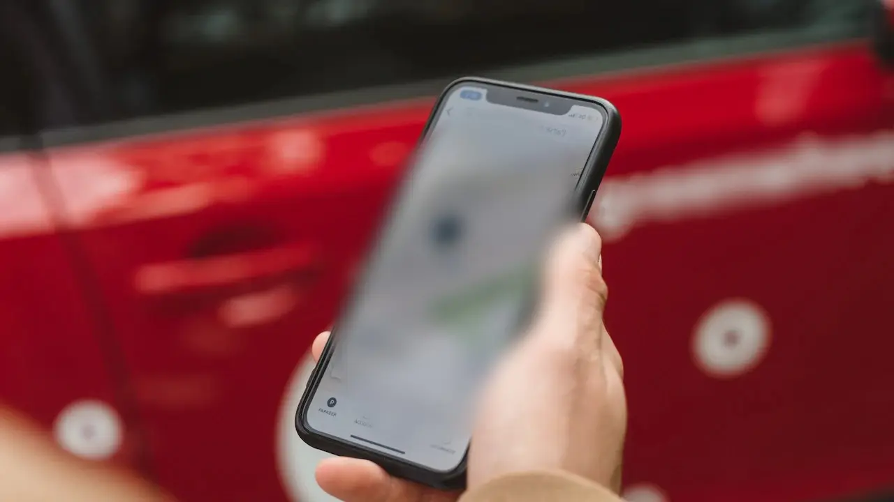 A hand holding a phone in front of a red car.