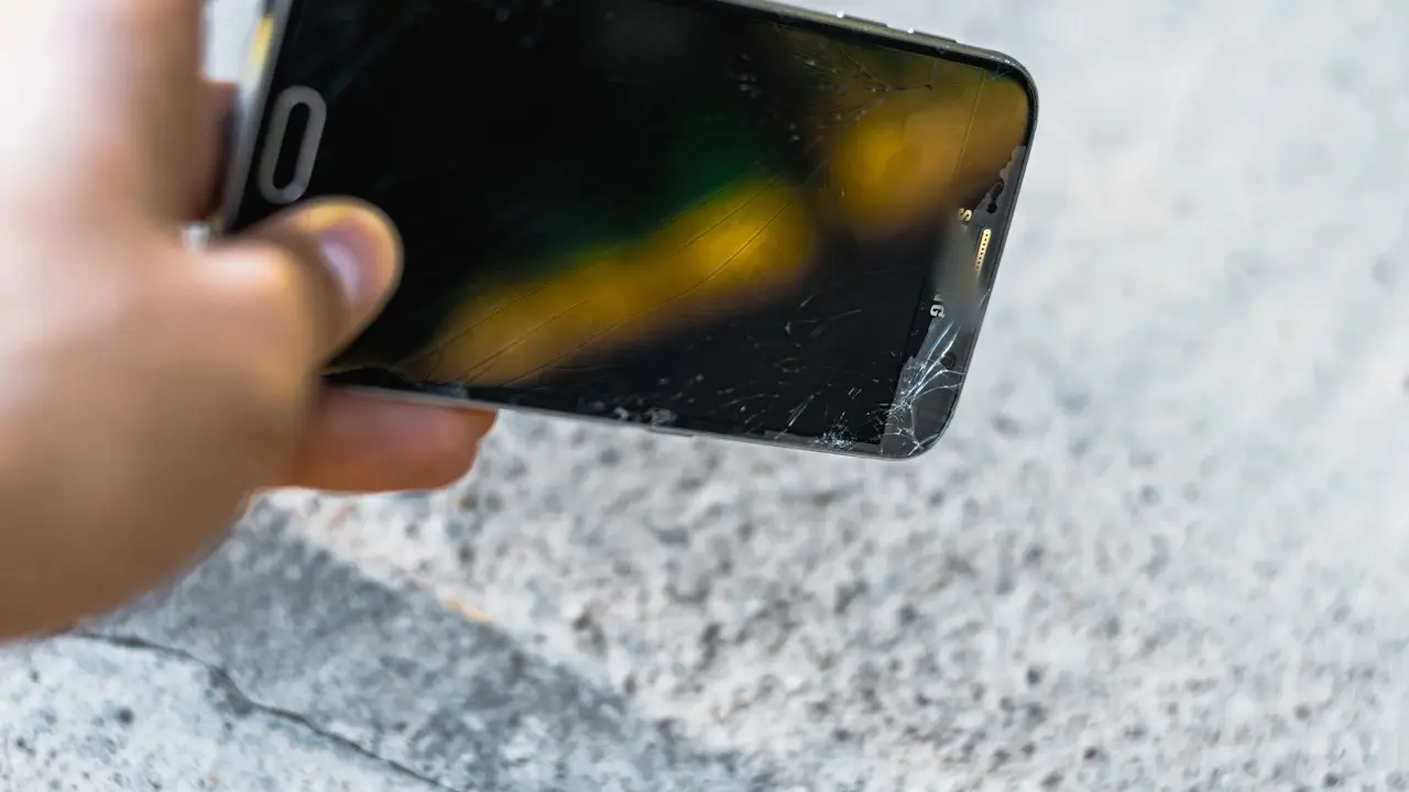 A hand holding a phone with a cracked corner over pavement.