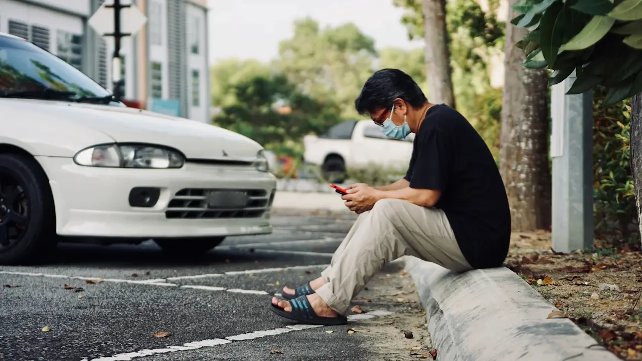 A man sitting on a kerb looking at his phone beside a parked white car.