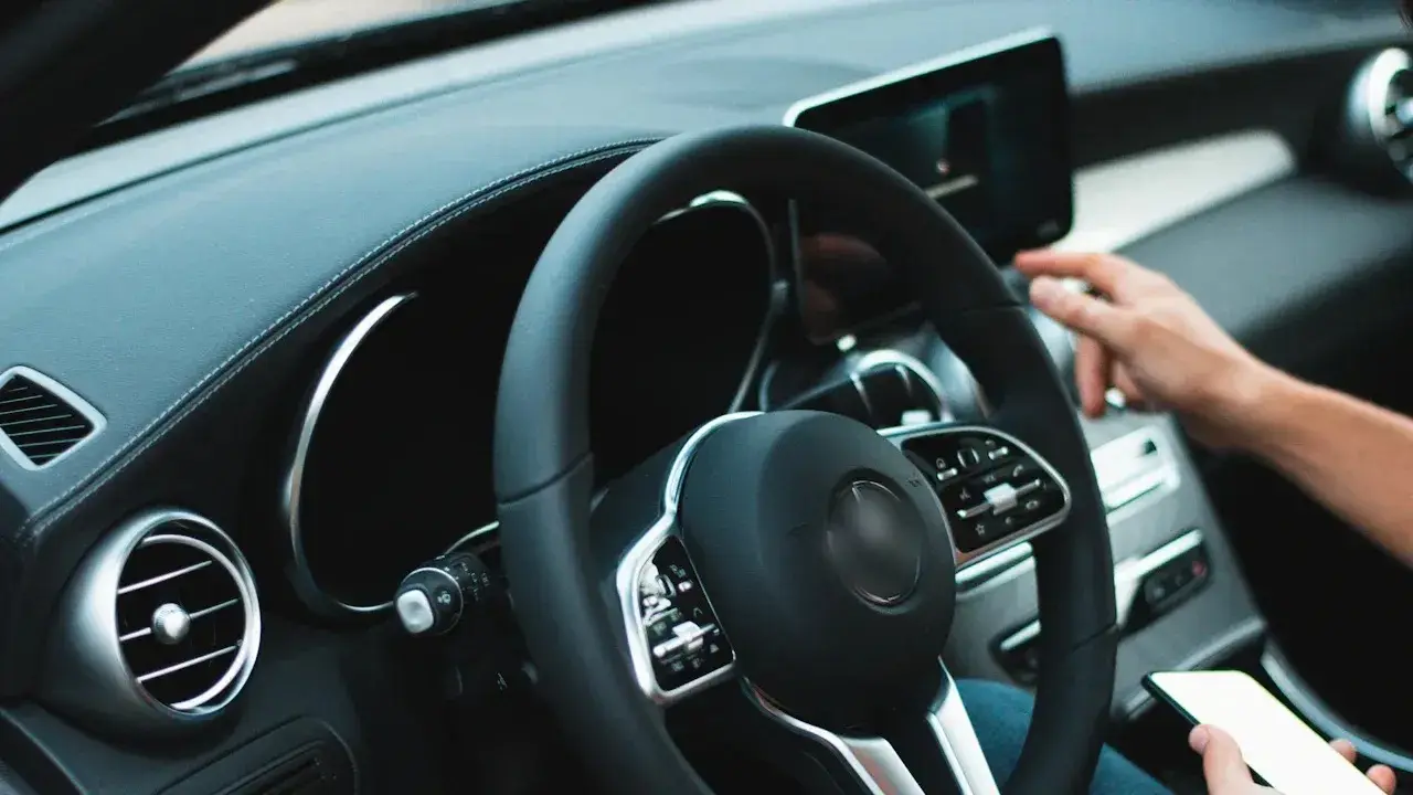 A driver's hand holding a smartphone below the steering wheel, the other hand on the dashboard touchscreen.