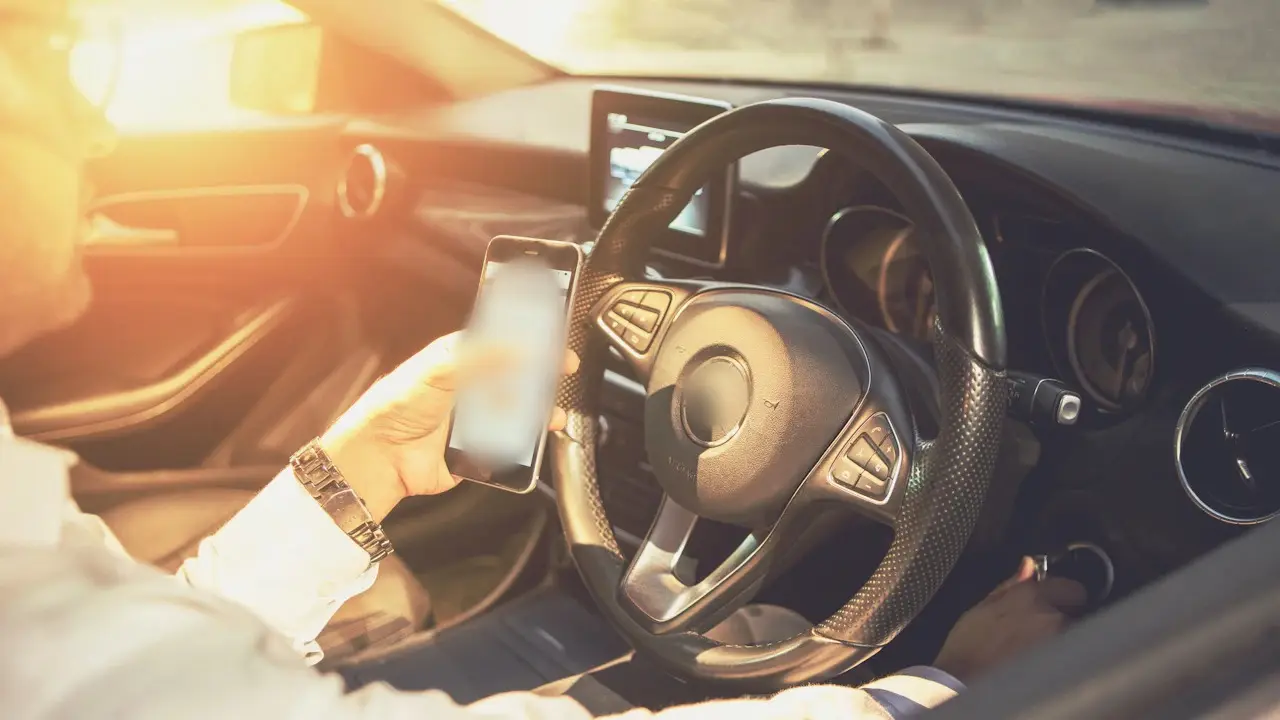 A hand holding a phone beside a car’s steering wheel.